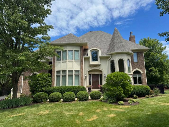 Large brick and stucco home with turret and landscaped front yard before exterior roofline work